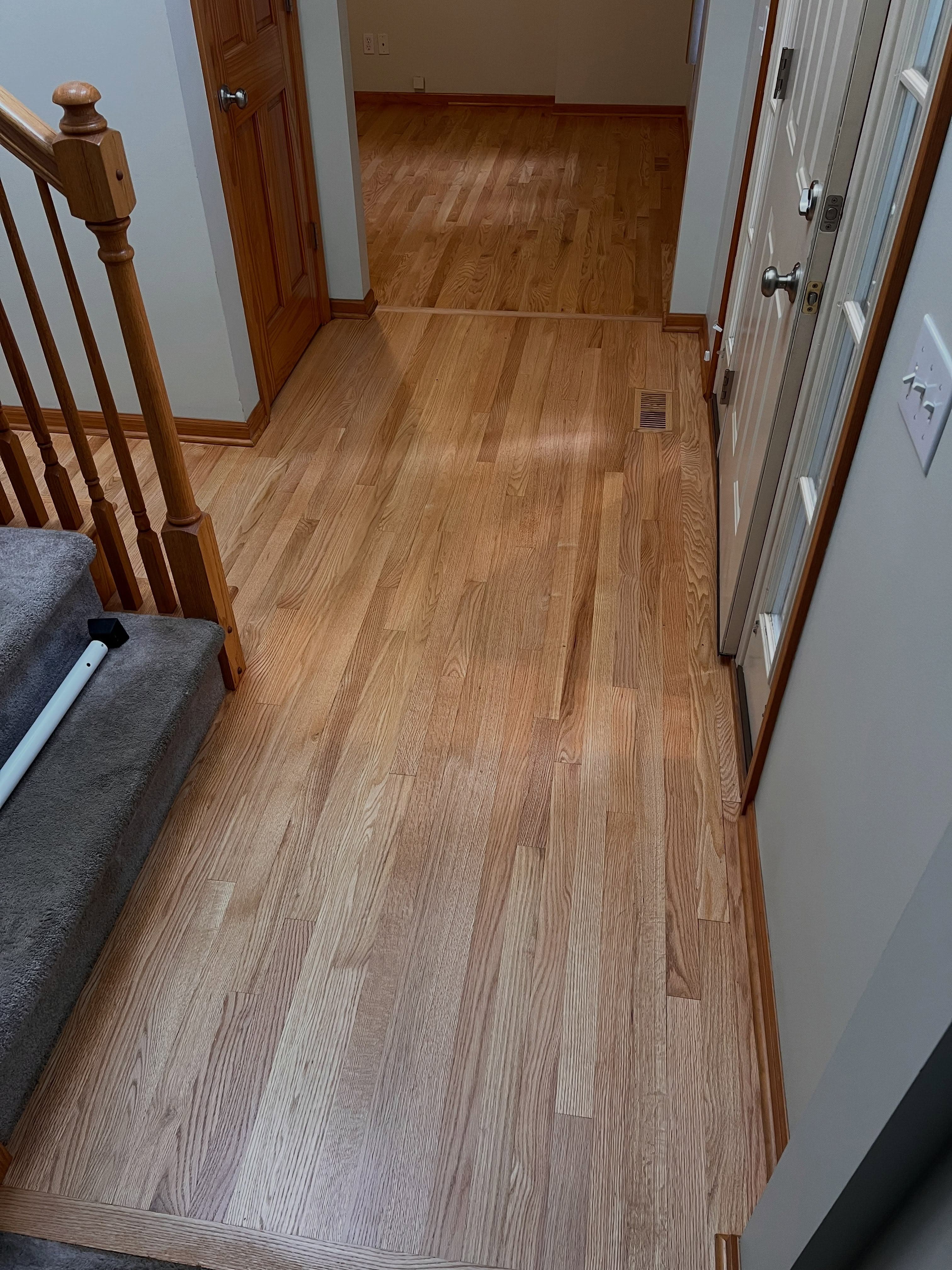Light wood plank flooring running through a residential hallway with wooden staircase and white trim